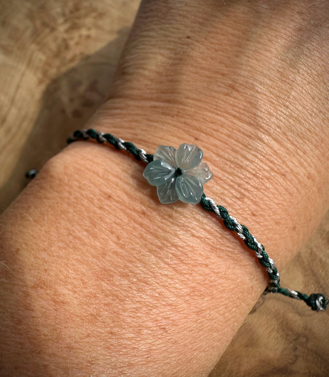 A close-up of a braided cord bracelet worn on a wrist, featuring a carved translucent gray-blue flower charm in the center.