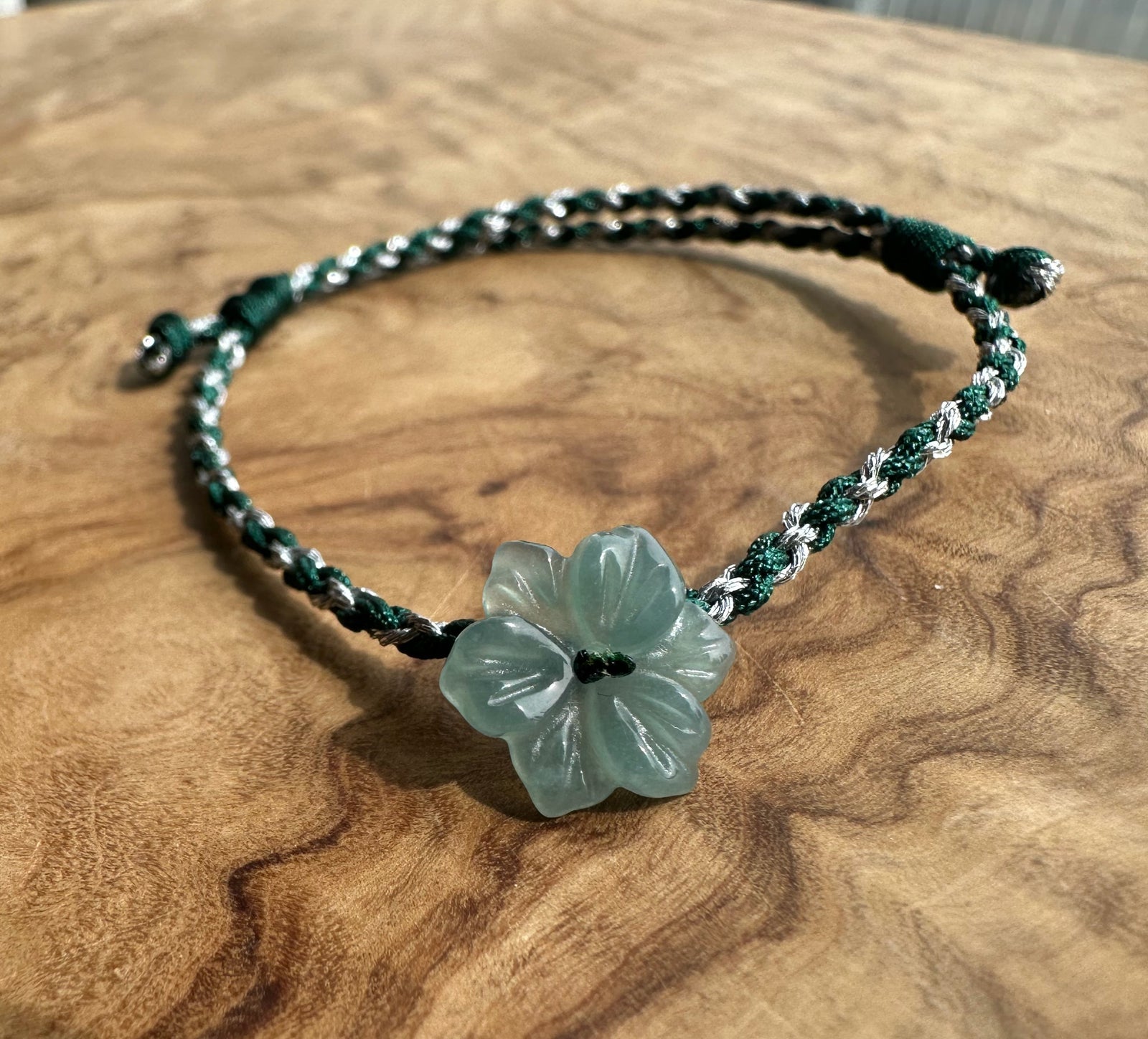 A braided cord bracelet with a carved light green flower charm, displayed on a textured wooden surface.