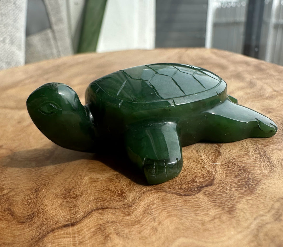 A polished dark green carved turtle figurine resting on a round wooden surface with a softly blurred indoor background.