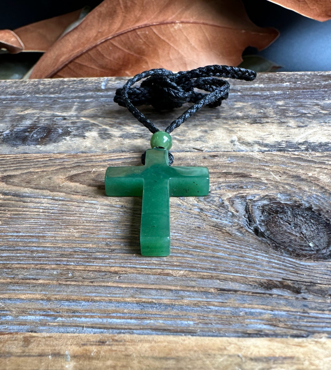 Dark green jade cross pendant with cord and round bead resting on weathered wood