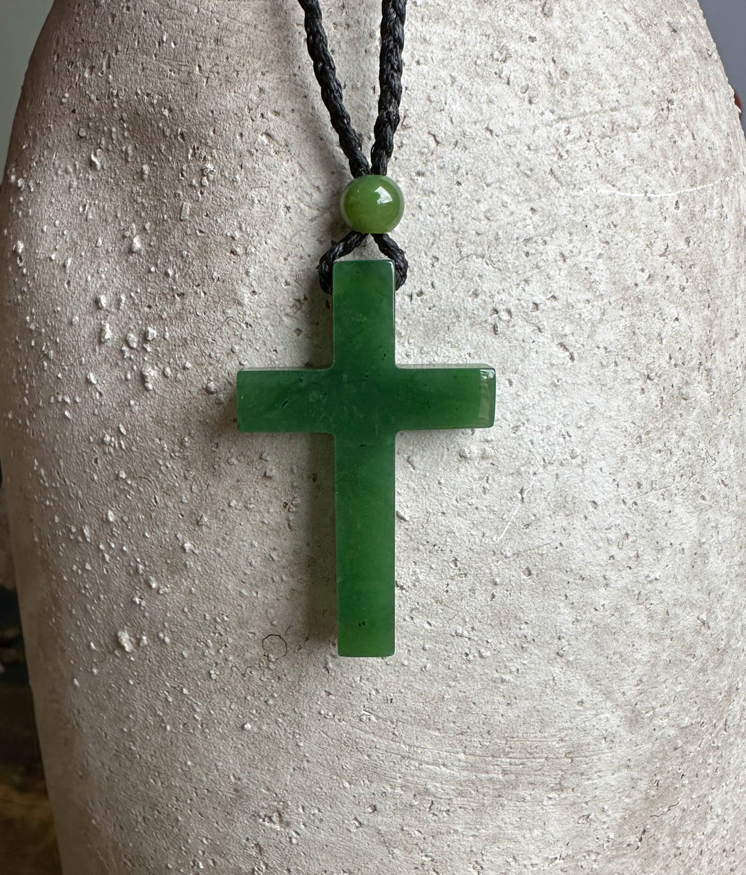 Dark green jade cross pendant on a black braided cord with a green bead above, displayed on a light textured surface