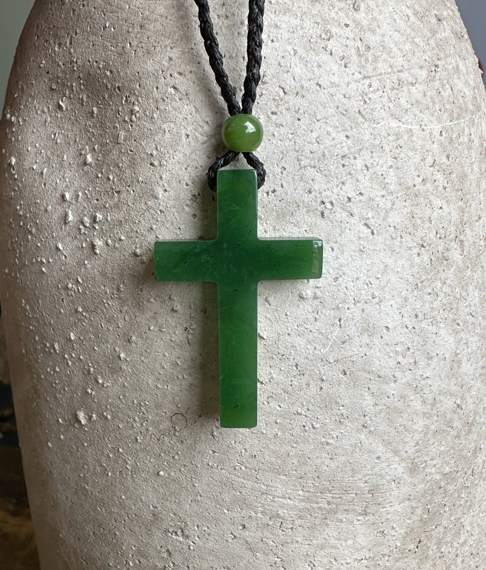 Dark green jade cross pendant on a black braided cord with a green bead above, displayed on a light textured surface