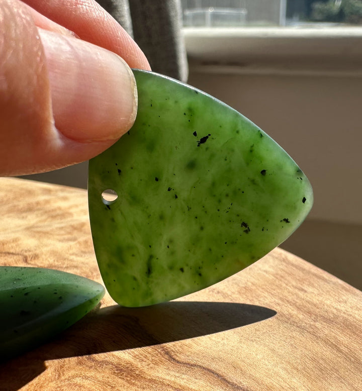 A hand holding a polished green jade teardrop-shaped pendant with a small hole, showing natural dark inclusions and a smooth glossy finish, set against a wooden table background.