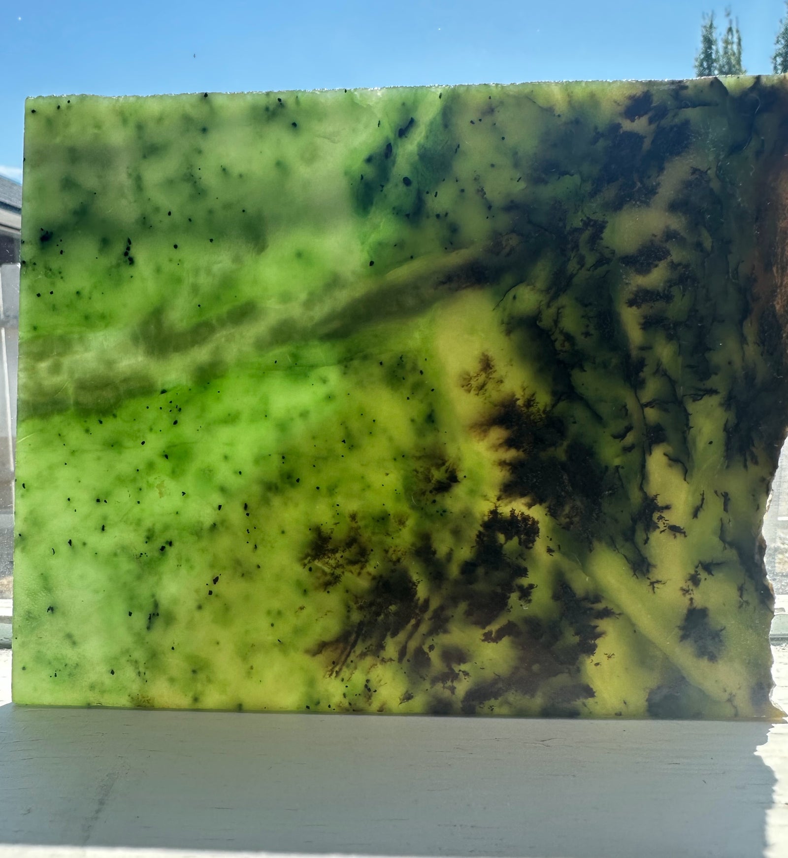 A rectangular slab of jade with a green, yellow, and black abstract pattern, photographed outdoors on a white surface with blue sky in the background.