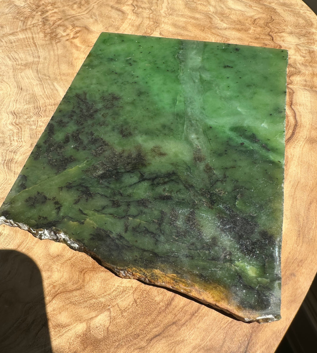 A green jade slab with natural marbling and rough edges, displayed on a light wood surface.