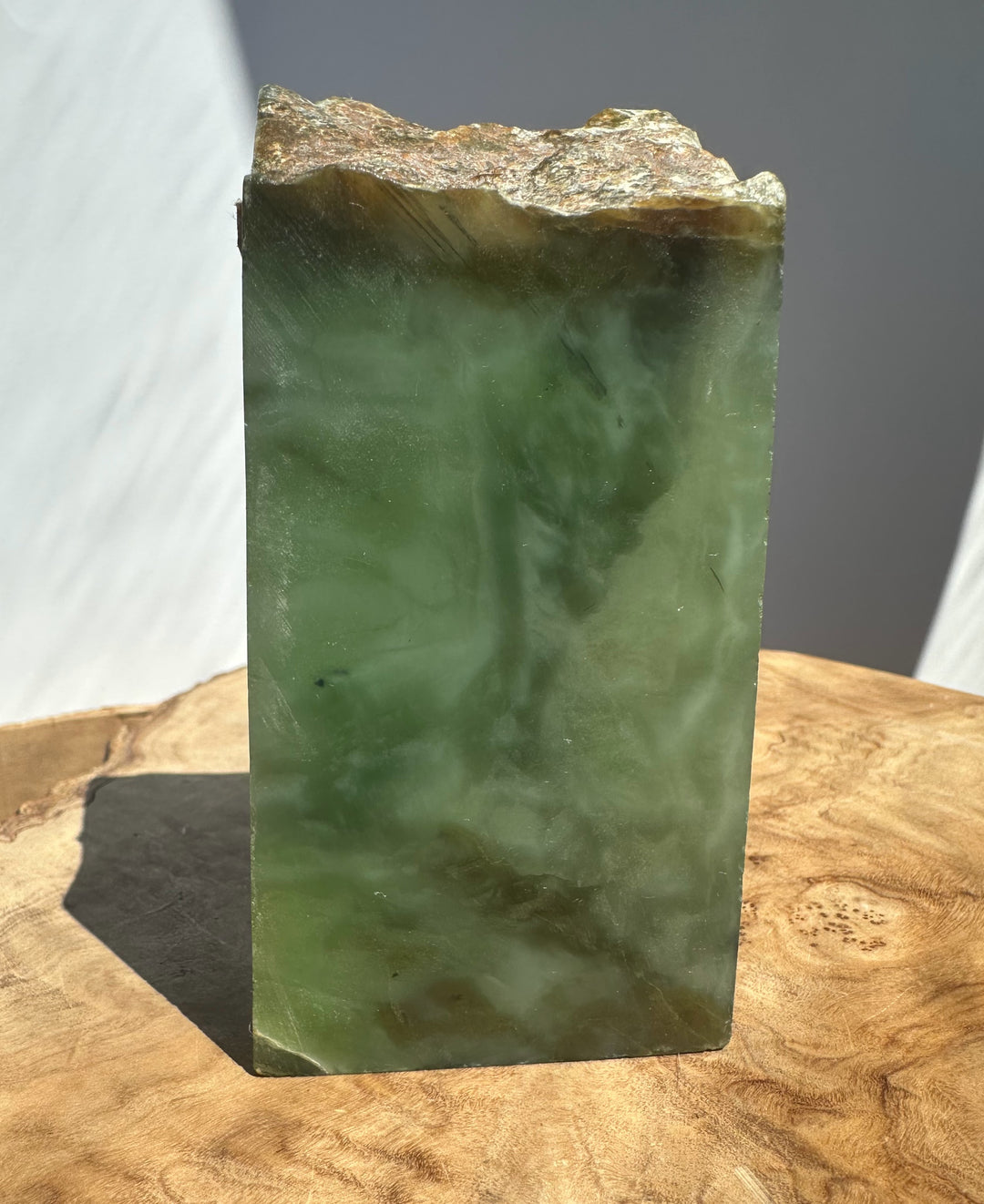 A rectangular green jade with a rough, uneven top surface, standing upright on a cut wood slice against a neutral gray background.