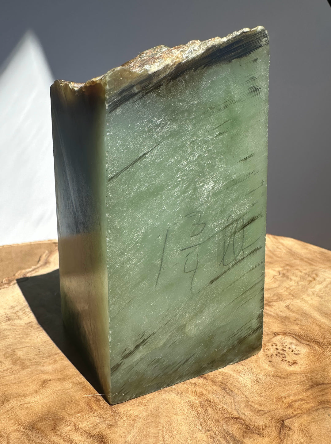 A rectangular jADE with a rough, uneven top edge, showing natural marbling in shades of green, gray, and brown. The slab has handwritten markings on the front face and is displayed on a wooden surface against a neutral gray background.