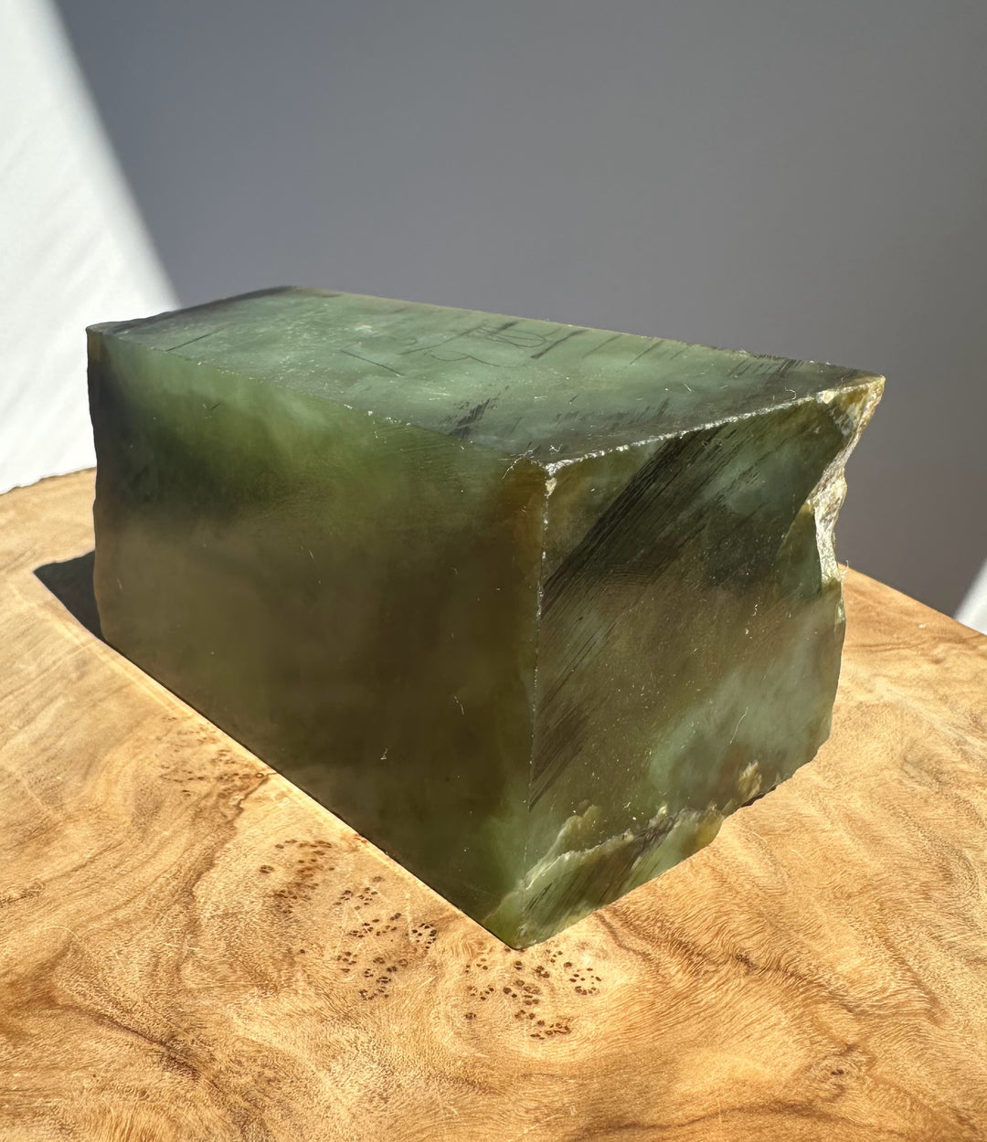 A rectangular green JADE block with rough, natural edges, displayed on a curved wooden surface against a neutral gray background.