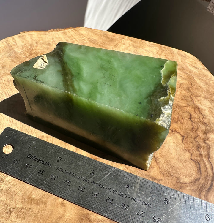A raw green jade block with rough edges, placed on a wooden surface next to a metal ruler for scale.