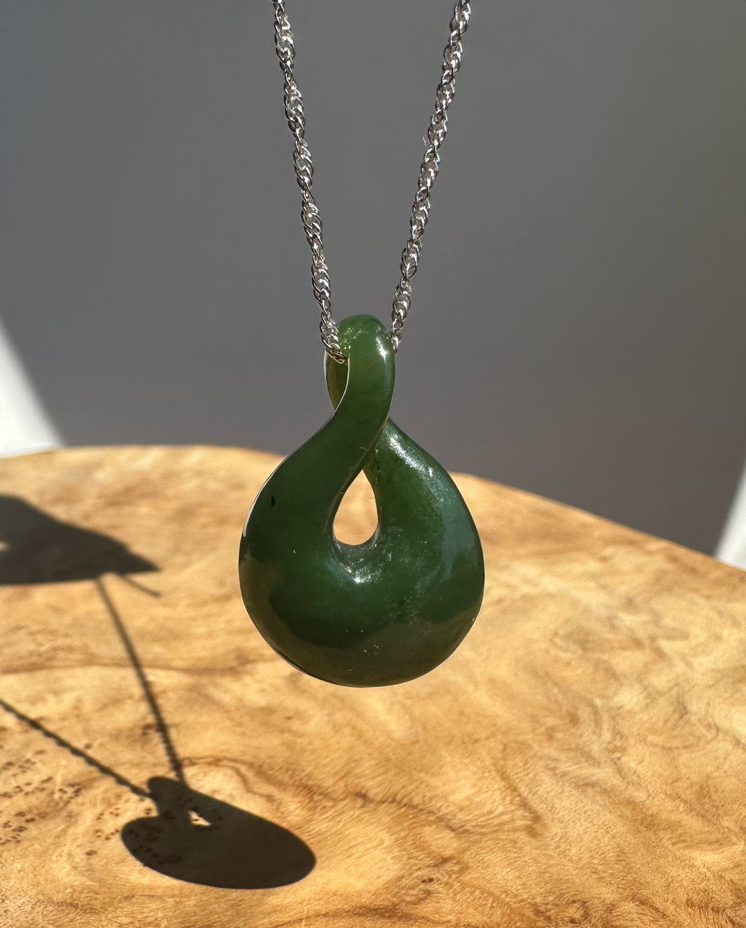 A polished dark green infinity pendant necklace on a silver chain, displayed on a wooden surface with a gray background.