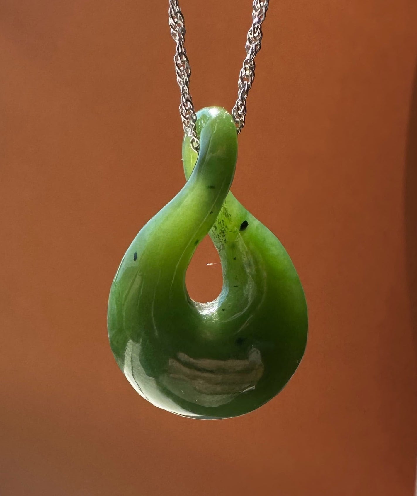 A green INFINITY-shaped pendant necklace with a silver chain, displayed against a warm brown background.