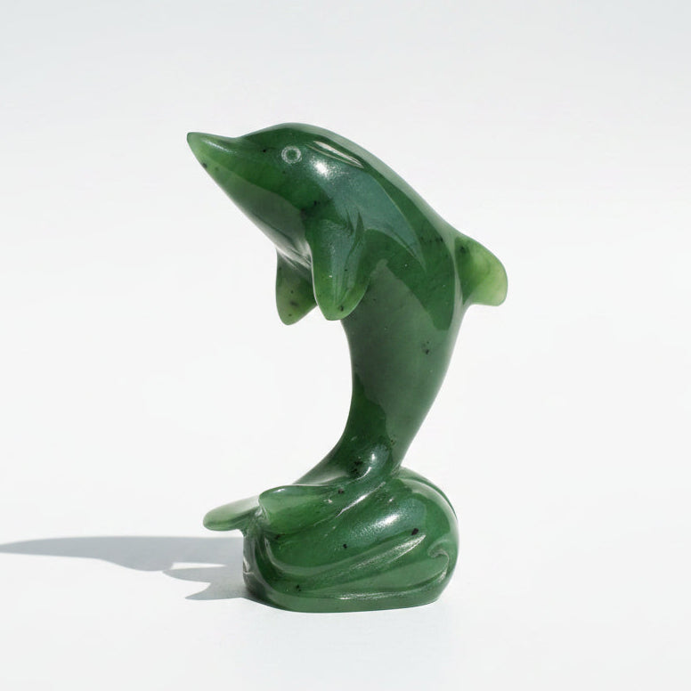 A polished green jade-like dolphin figurine standing on a small base, photographed on a wooden surface against a light neutral backdrop.
