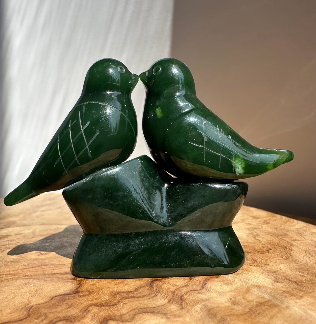 A dark green polished stone sculpture of two kissing birds perched on a matching base, displayed on a wooden surface with a neutral background.