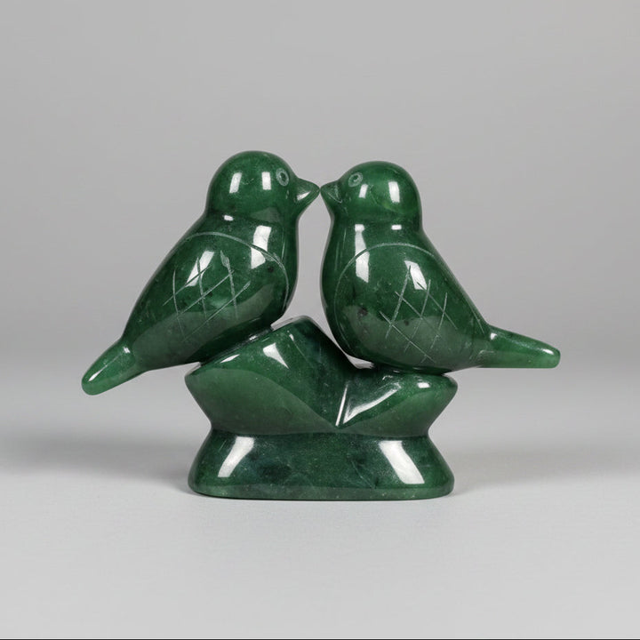 A green jade sculpture of two small birds perched on a matching base, photographed on a grey background 