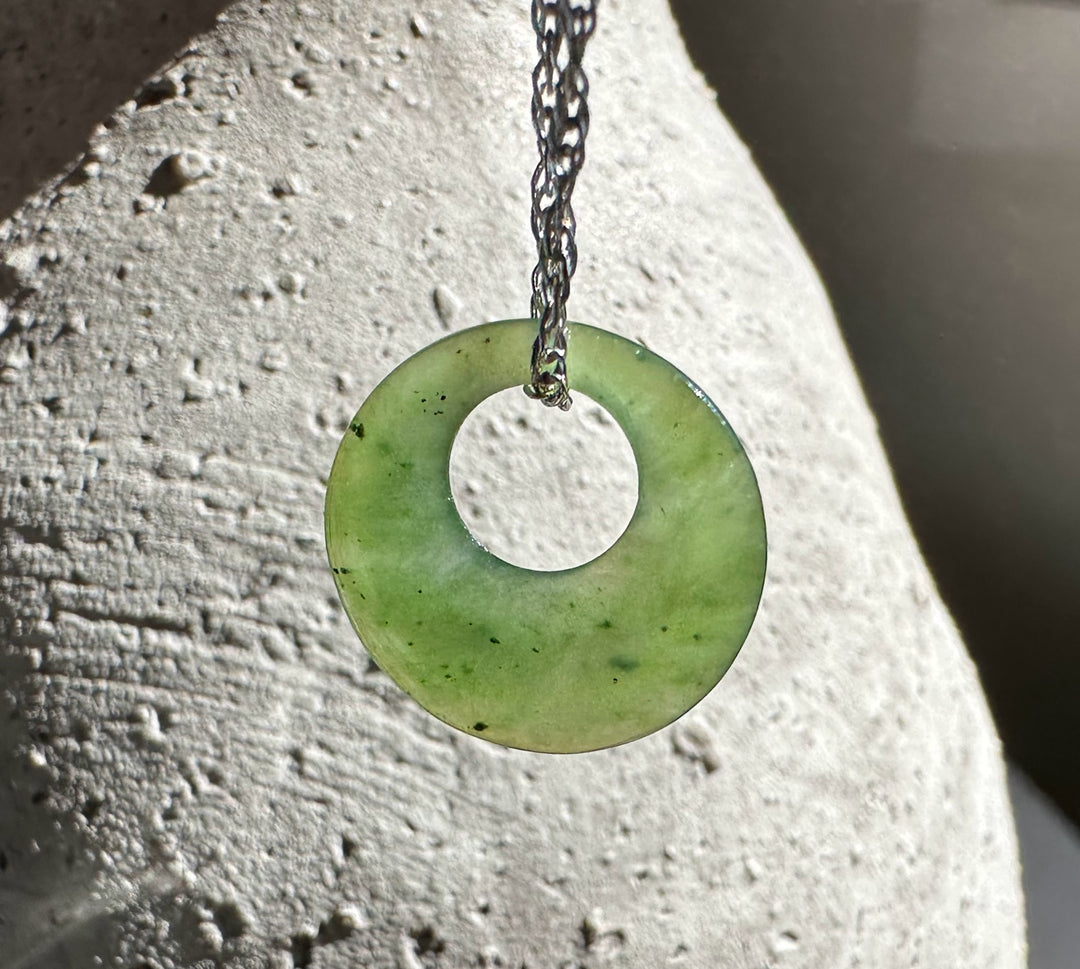 Pale green jade donut pendant hanging on a dark chain against a light textured stone surface