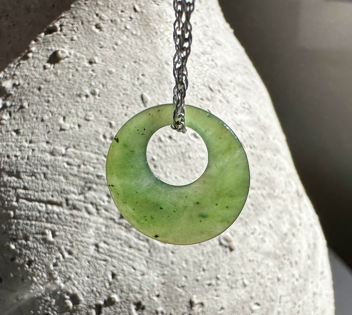 Pale green jade donut pendant hanging on a dark chain against a light textured stone surface