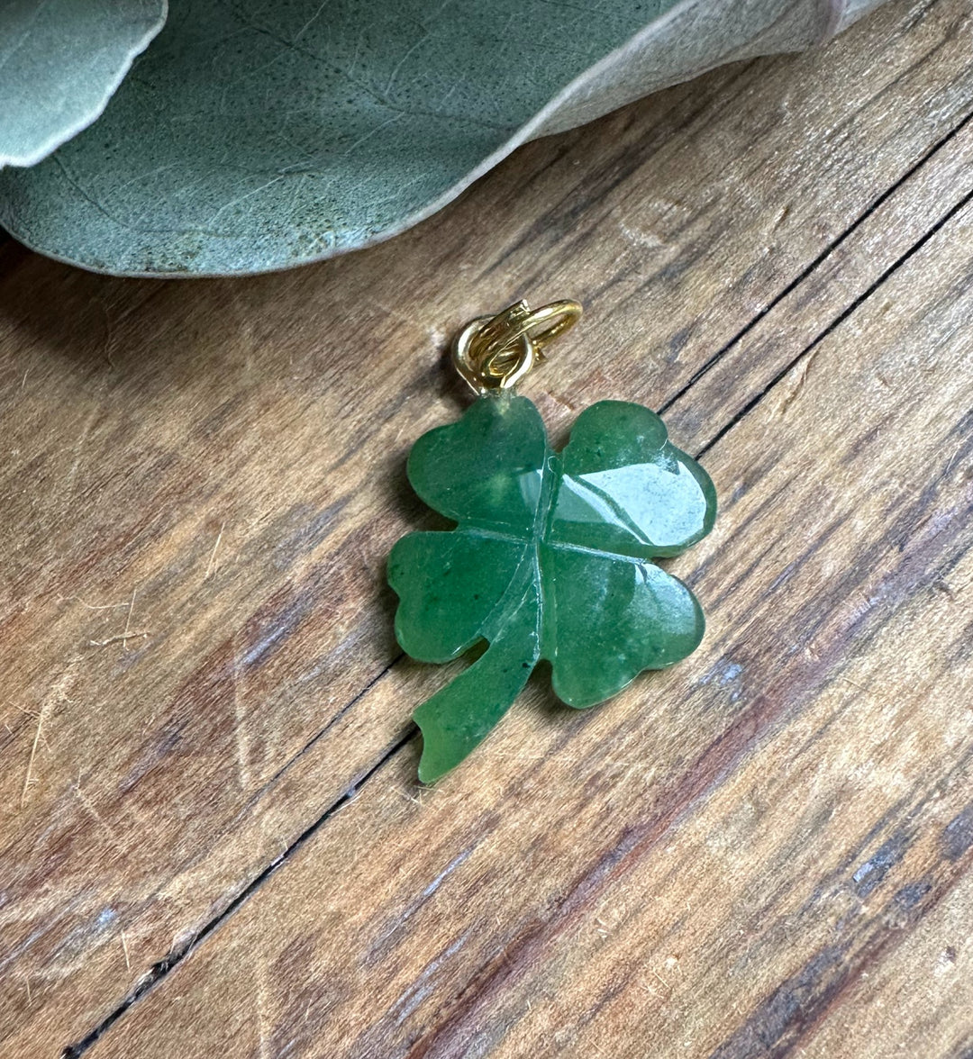 Green shamrock charm with a gold loop resting on wood beside a eucalyptus leaf.