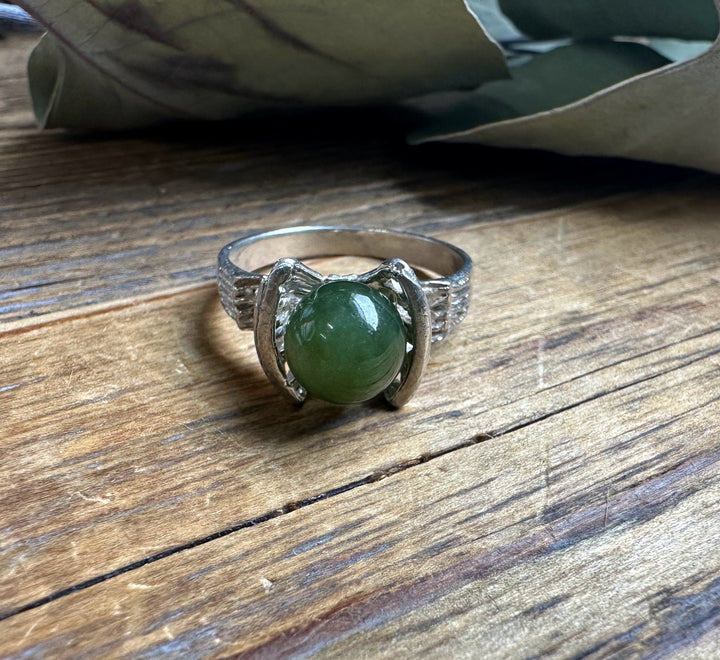 Silver ring on a wooden surface featuring a large green jade bead cradled by curved side settings.