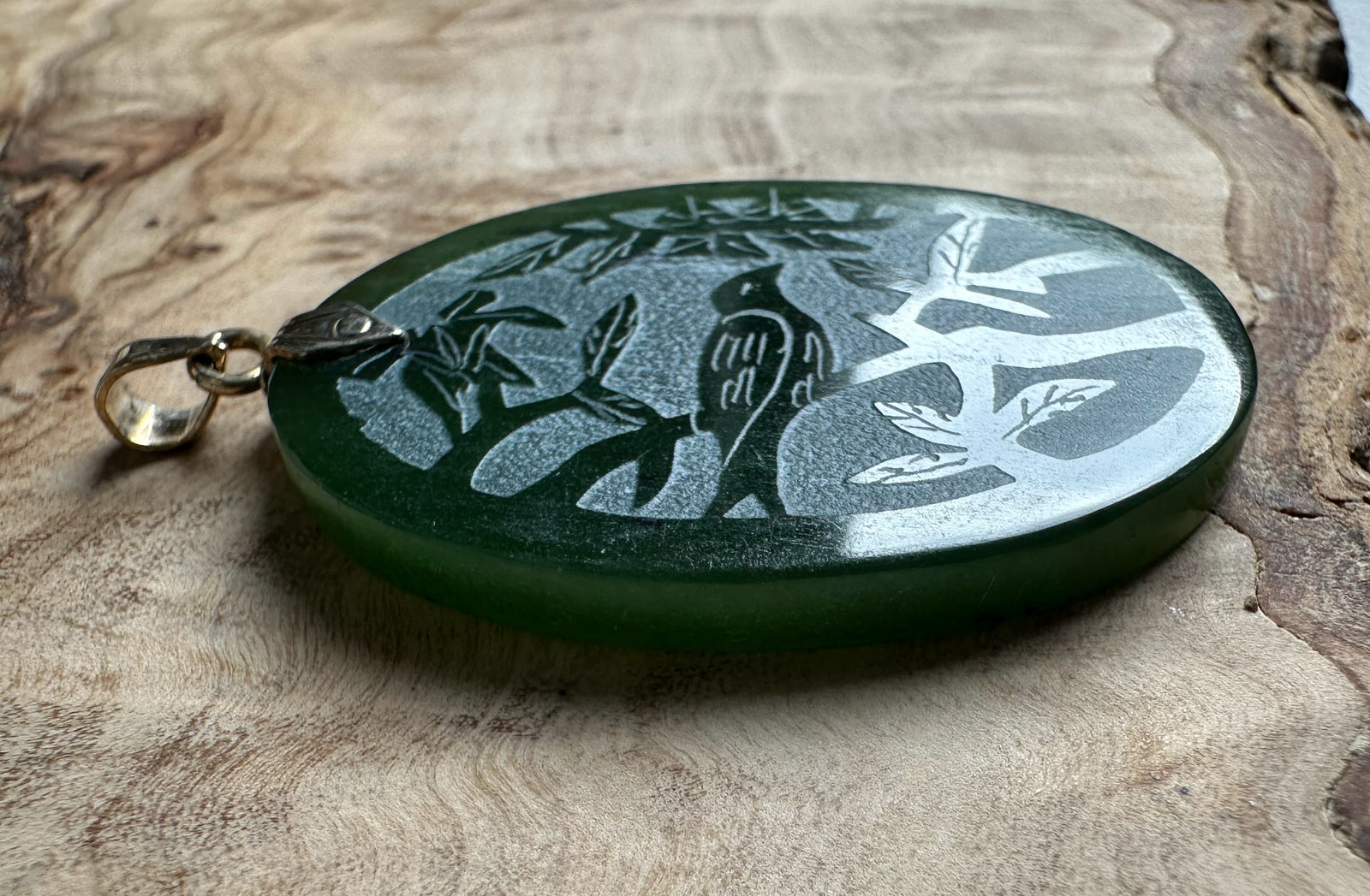 Oval green jade pendant with an etched bird and bamboo scene and a silver bail on a wooden surface