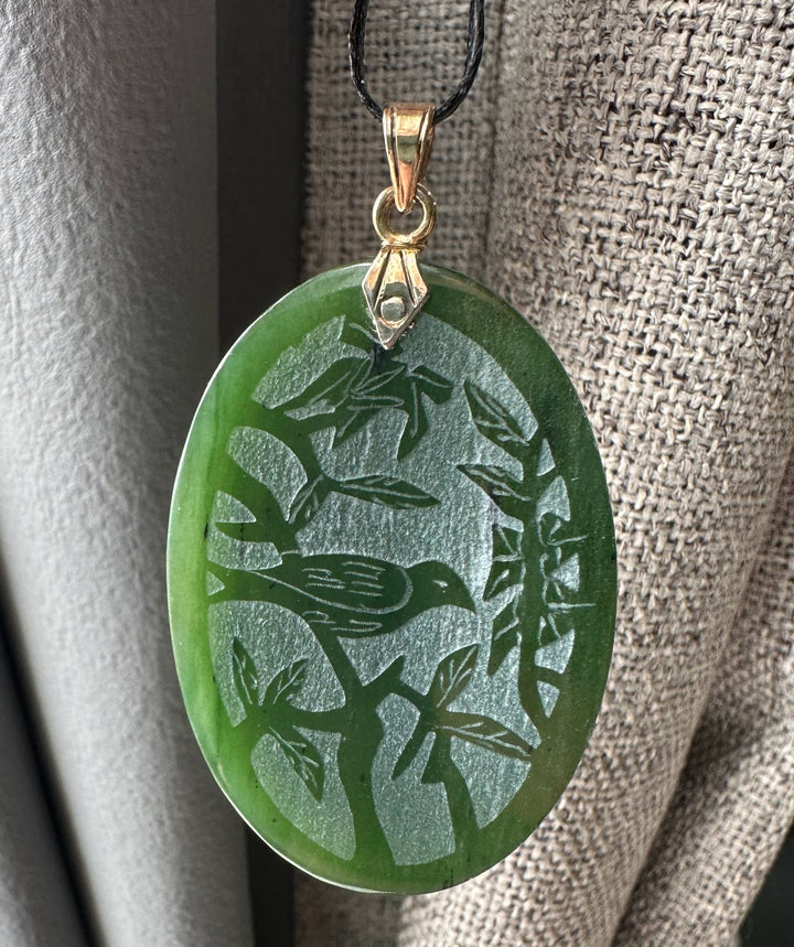 Oval green jade pendant featuring a bird among leaves with a gold bail on a black cord.