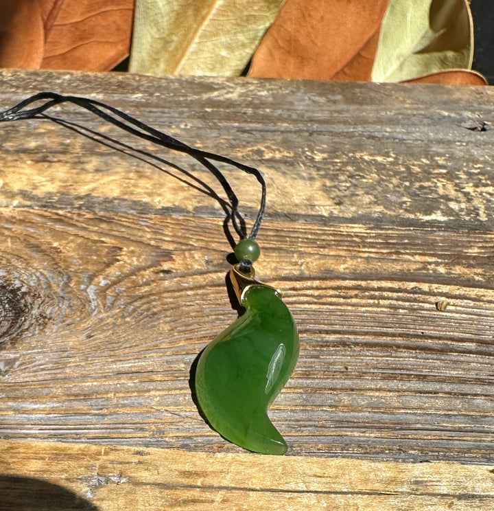 Green nephrite jade pendant with gold-tone bail on a black cord resting on a wooden surface