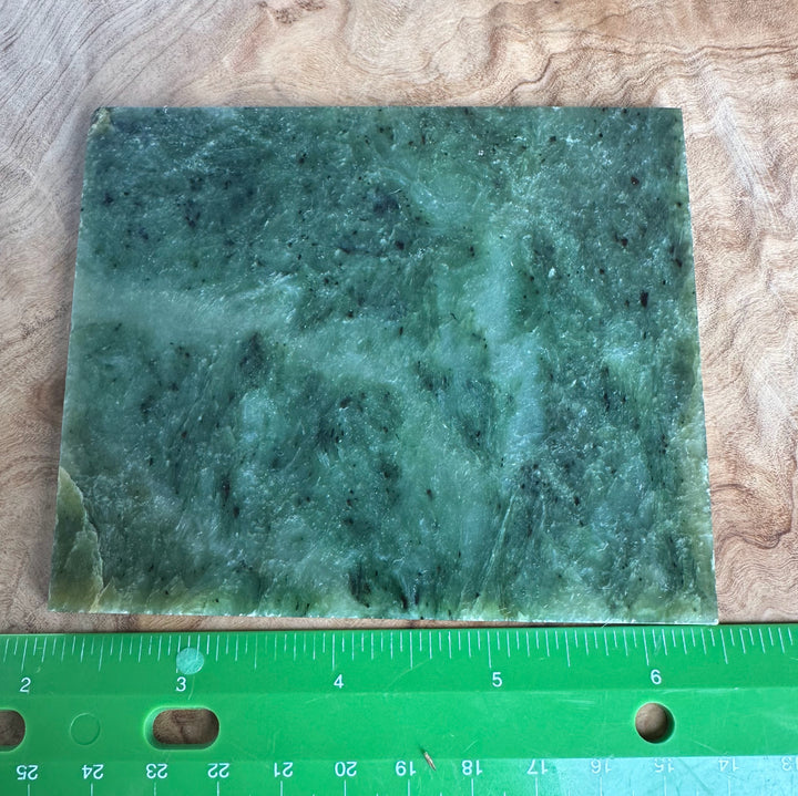 A rectangular green stone slab with natural marbled texture and dark inclusions, shown on a wood surface with a green ruler along the bottom edge for scale.