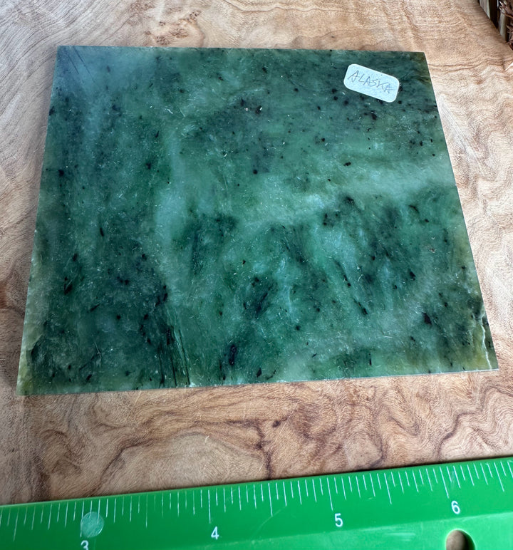 A square green jade slab with natural marbling and dark inclusions, placed on a wooden surface with a green ruler at the bottom for scale.
