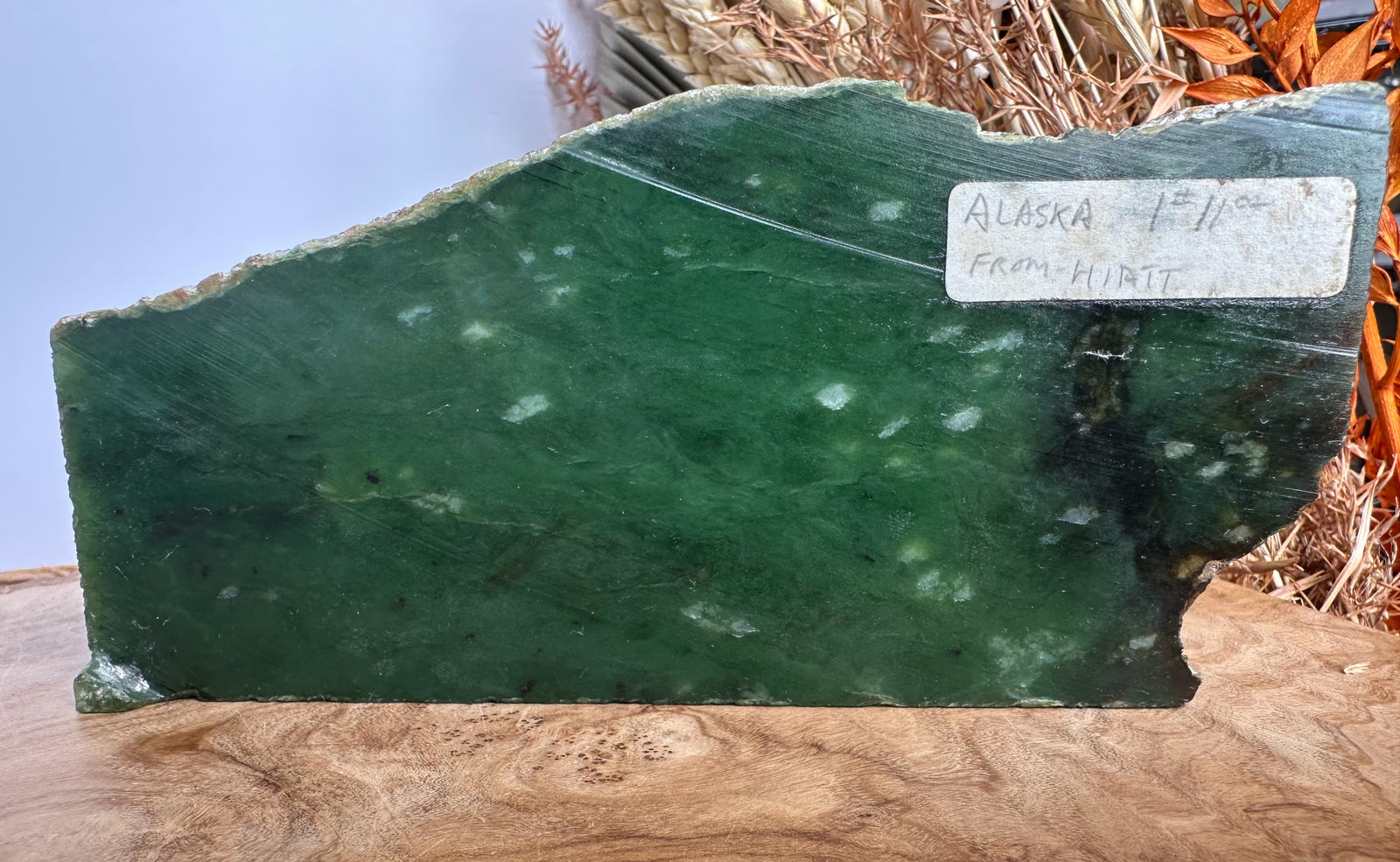 A raw, polished green stone slab with natural jagged edges, featuring white inclusions and a handwritten white label reading 'ALASKA 1/4 FROM HIATT'. The slab is displayed on a wooden surface with a light blue background and dried brown grass behind it.