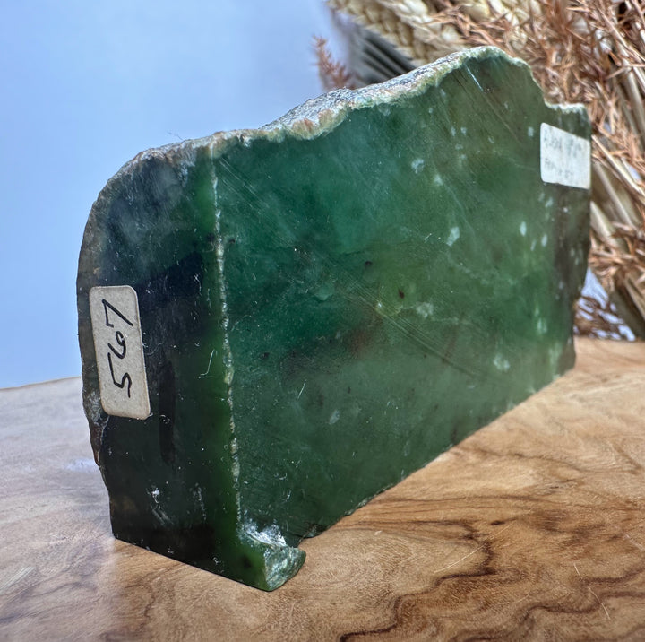 A green jade stone slab with a rough, natural cut edge, resting on a wooden surface. The slab has two white paper labels with handwritten numbers, and a dried plant is visible in the background against a blue backdrop.