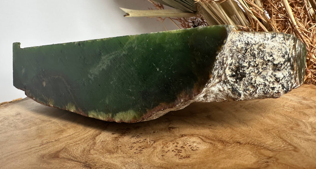 A raw green jade stone slab with a rough, textured white and brown outer crust, resting on a wooden surface with dried grass in the background.