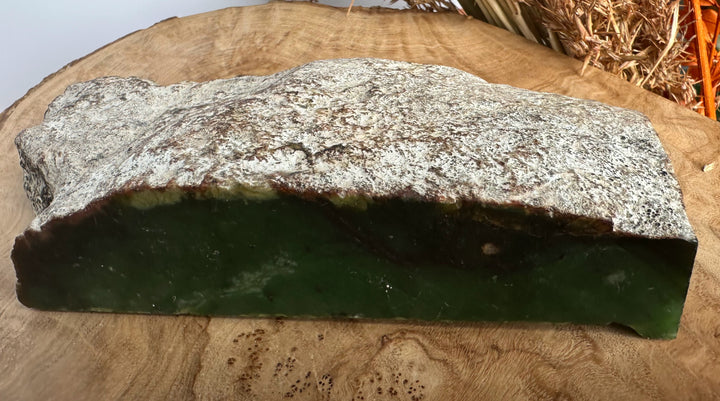A raw JADE slab with a dark green base and a heavily textured, speckled gray-white top, displayed on a rustic wooden slice.
