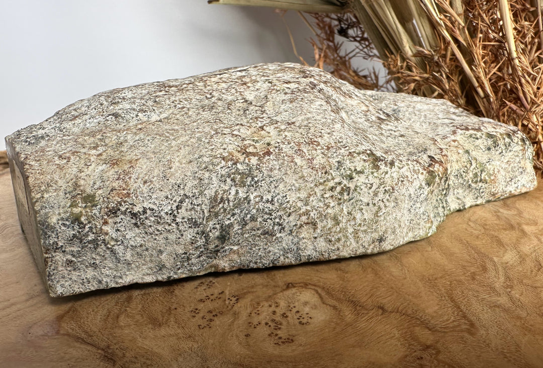 A large natural rough stone slab with a speckled granite-like texture in gray, beige, and brown tones, displayed on a wooden surface with dried grass in the background.