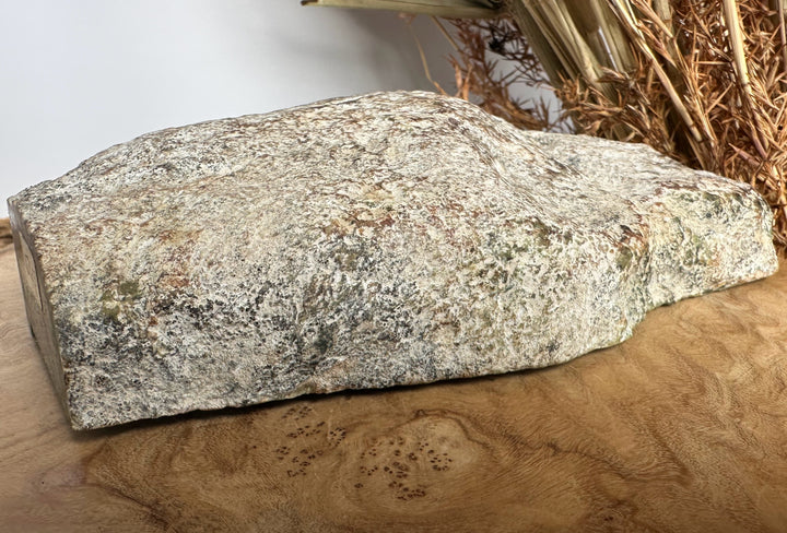 A large natural rough stone slab with a speckled granite-like texture in gray, beige, and brown tones, displayed on a wooden surface with dried grass in the background.