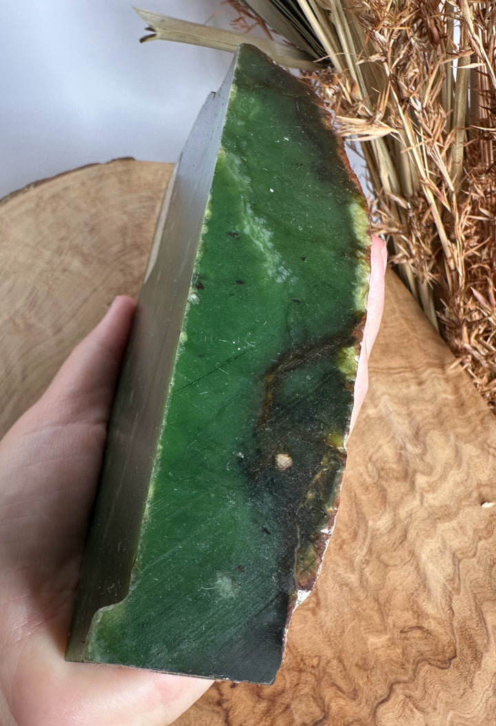 A hand holding a thick, green stone or jade rectangular slab with natural dark veining, set against a rustic wood and dried grass background.