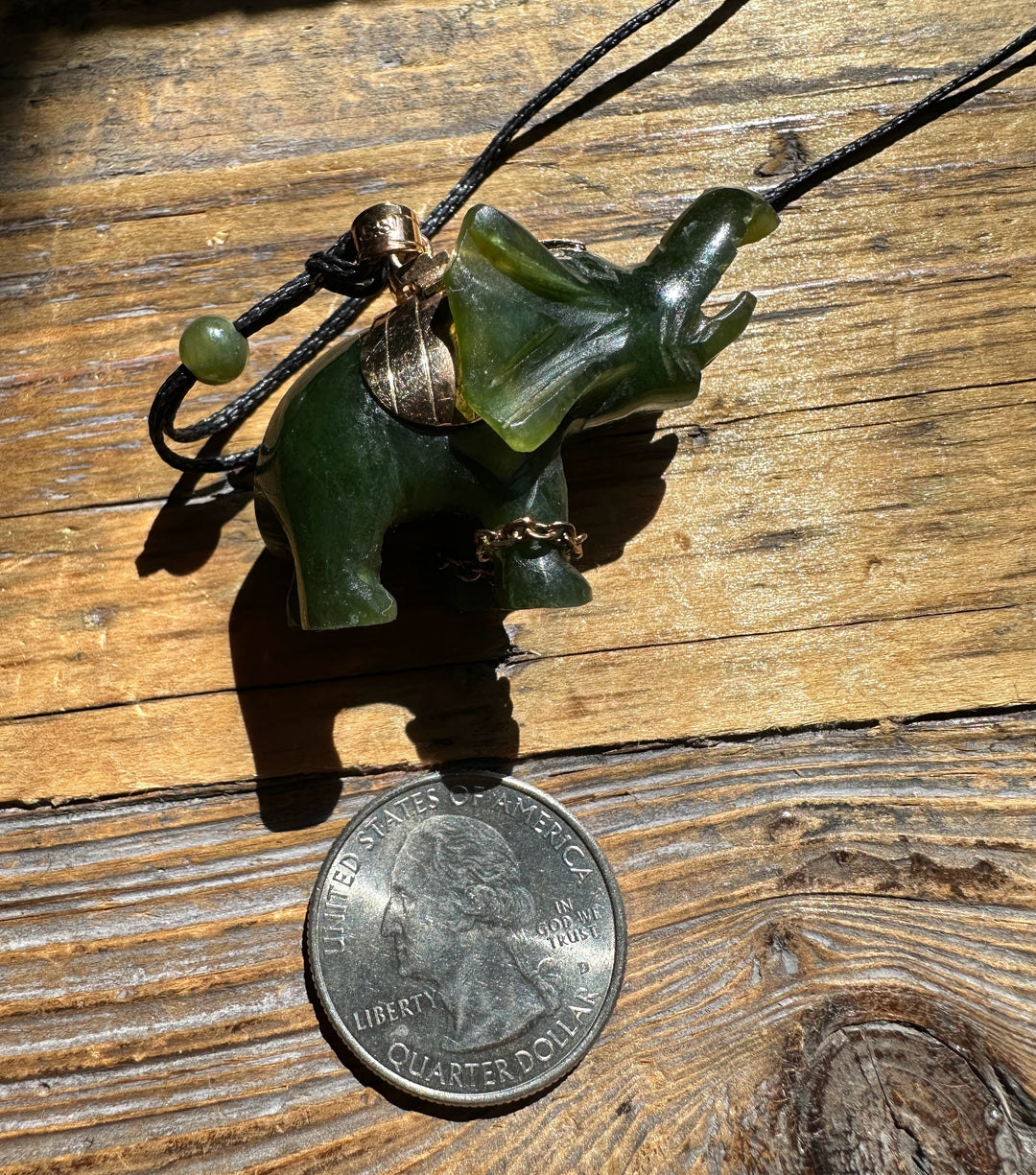 Jade elephant pendant with gold accents on black cord laid on wood beside a quarter for scale
