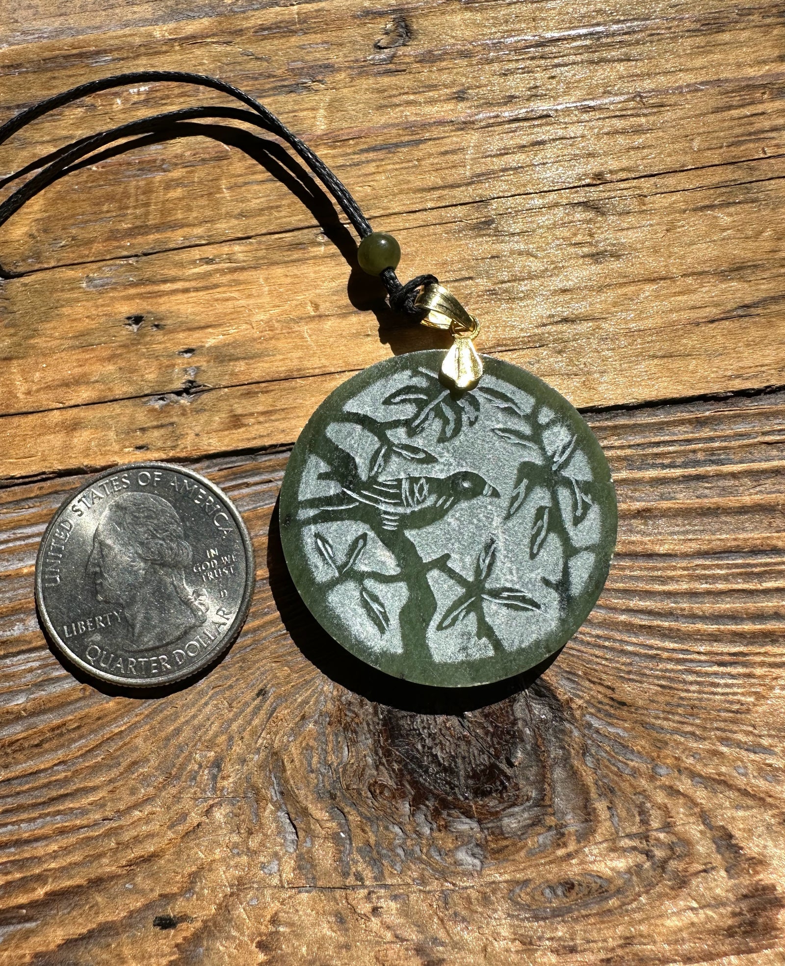 Round green jade pendant carved with a bird among leafy branches, hanging on a black cord with a gold bail