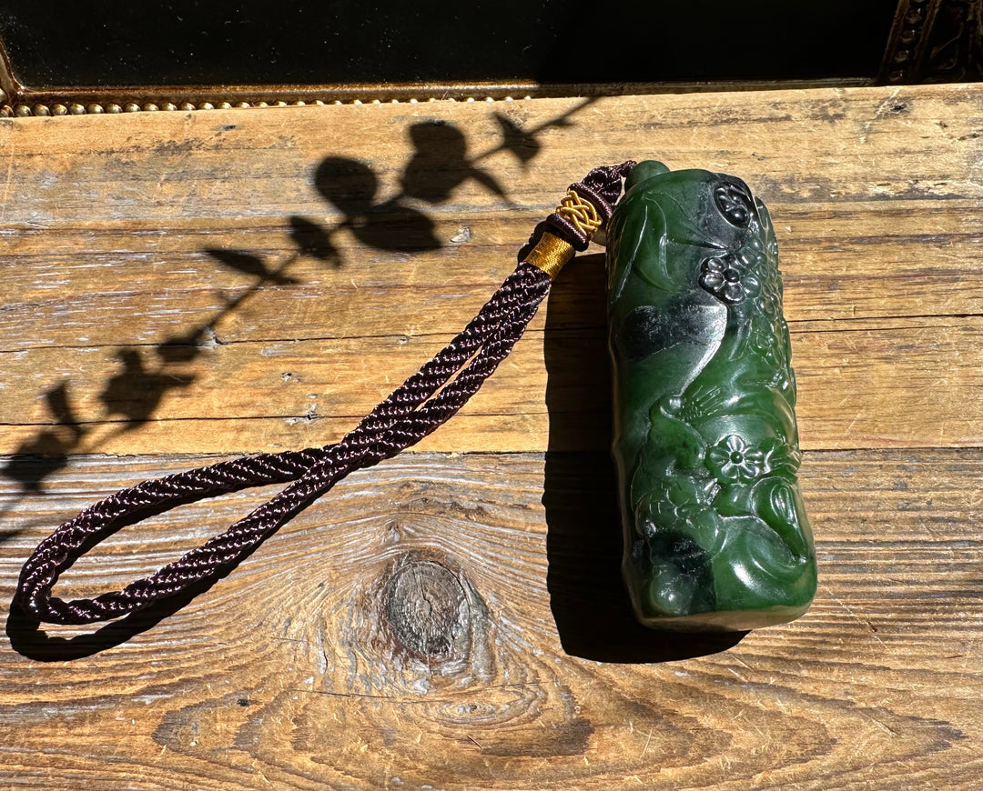 Green jade hand held pendant with floral carvings and a brown braided tassel shown on a wooden surface