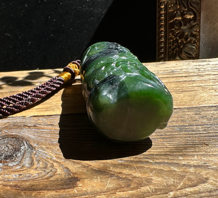 Green jade pendant with floral carving resting on wood, attached to a brown braided cord