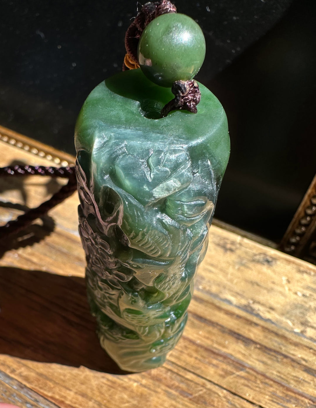 Green jade pendant with intricate carvings and a round top bead on a brown cord.