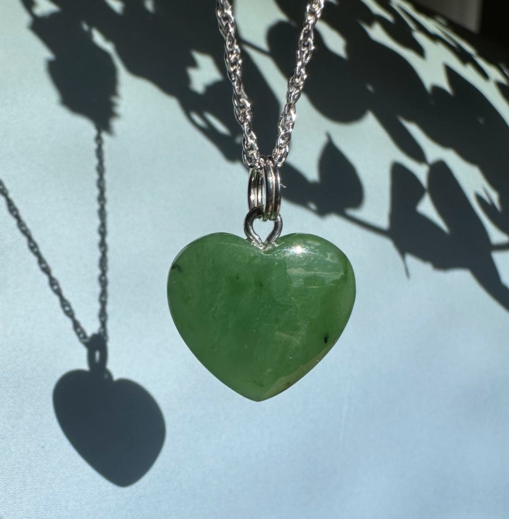 Green jade heart pendant hanging from a silver chain against a pale blue background