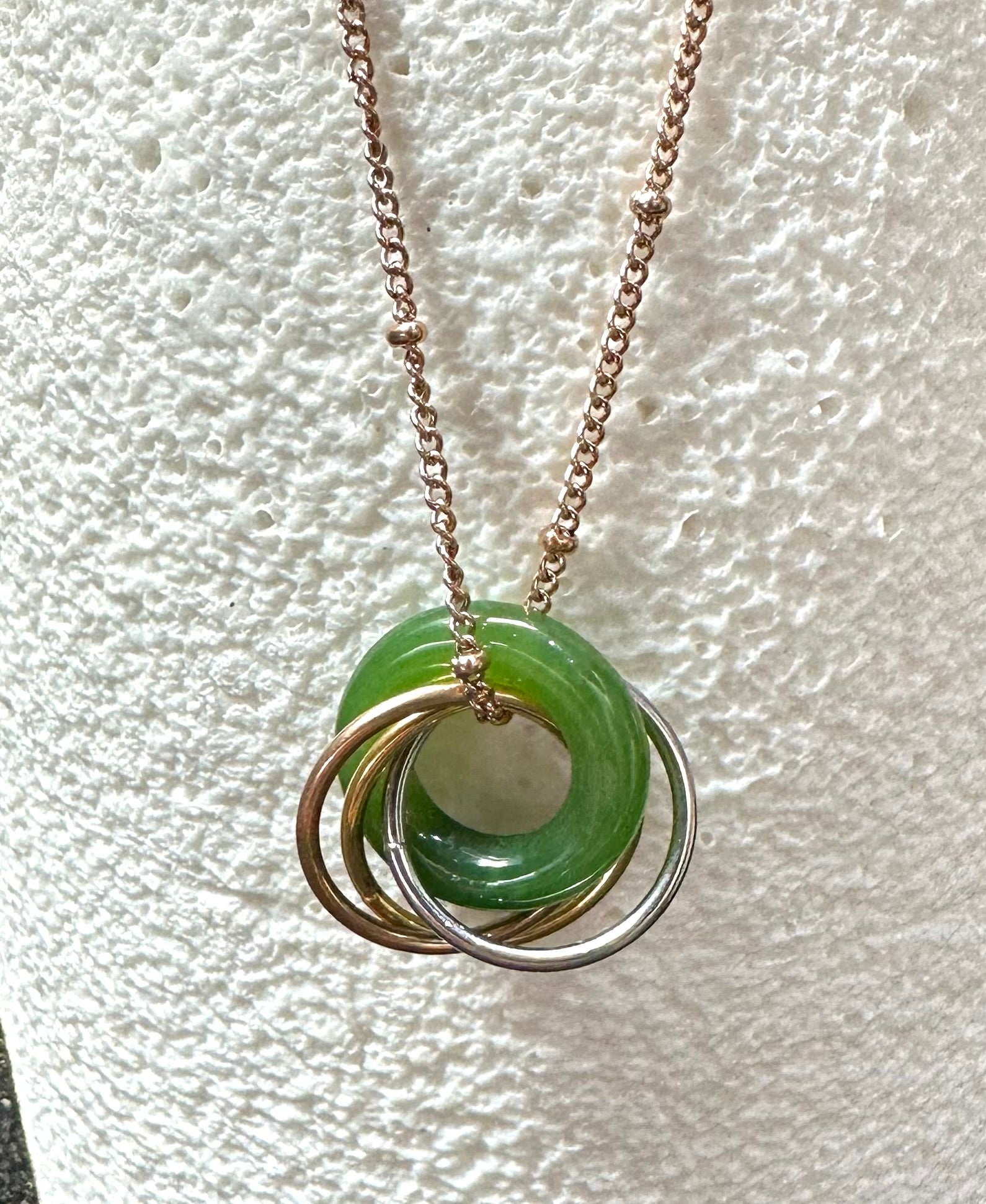 Close up of green jade pendant showing overlapping rings in gold copper and silver tones on a rose gold chain