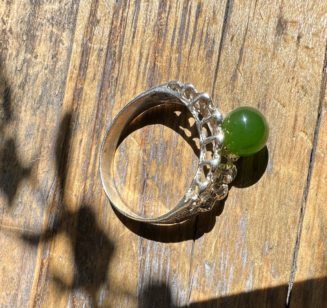 Silver ring with a round green jade cabochon and intricate openwork shoulders on a wooden surface.