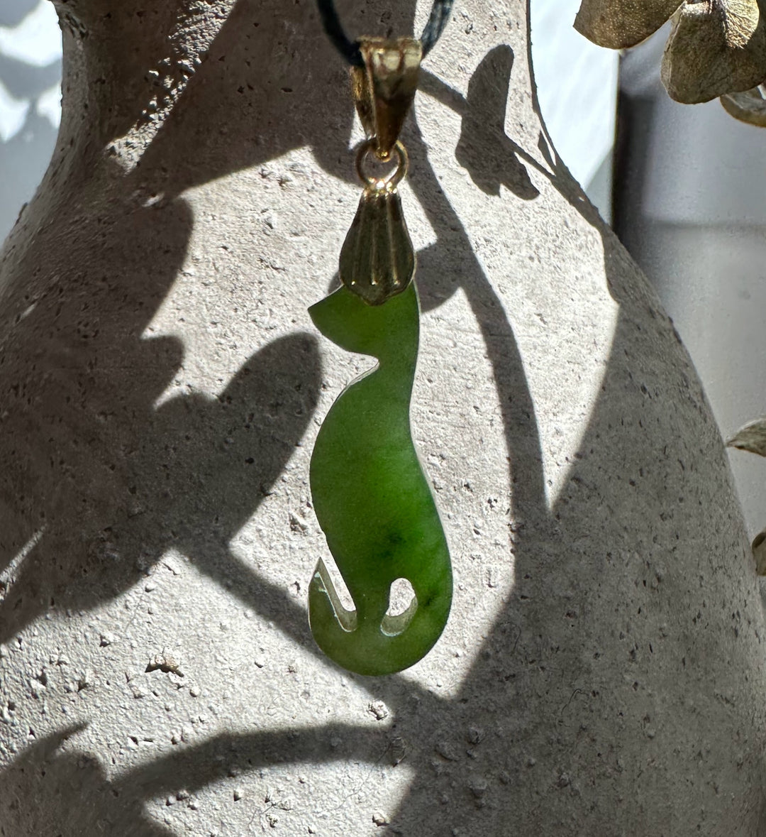 Close up of a green jade cat pendant with a gold bail against a pale stone surface