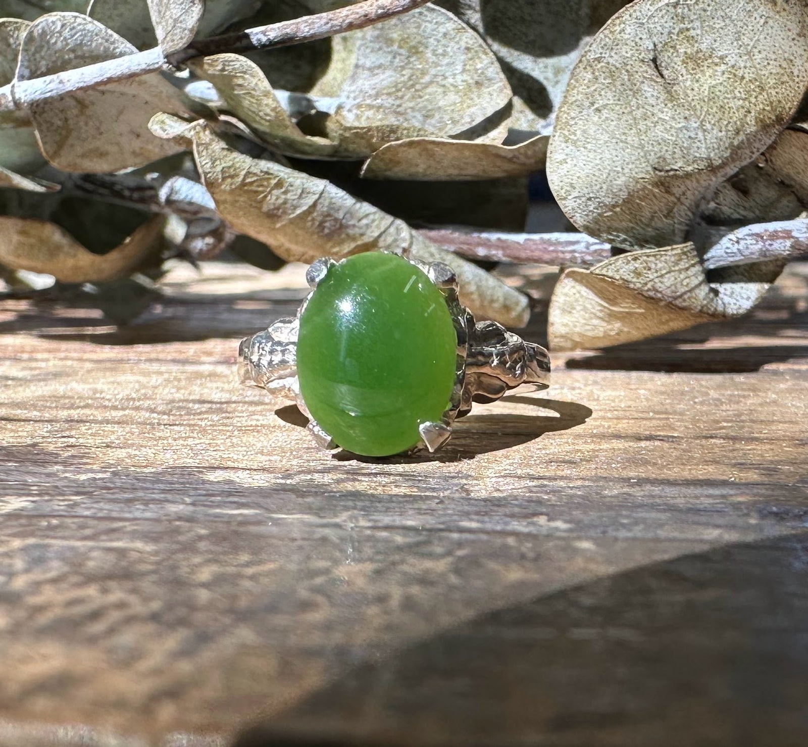 Close-up of the jade ring on a wooden surface with dried leaves in the background, highlighting the green jade and silver setting