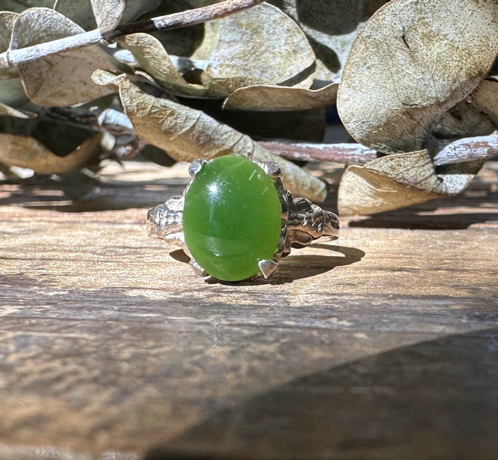 Close-up of the jade ring on a wooden surface with dried leaves in the background, highlighting the green jade and silver setting