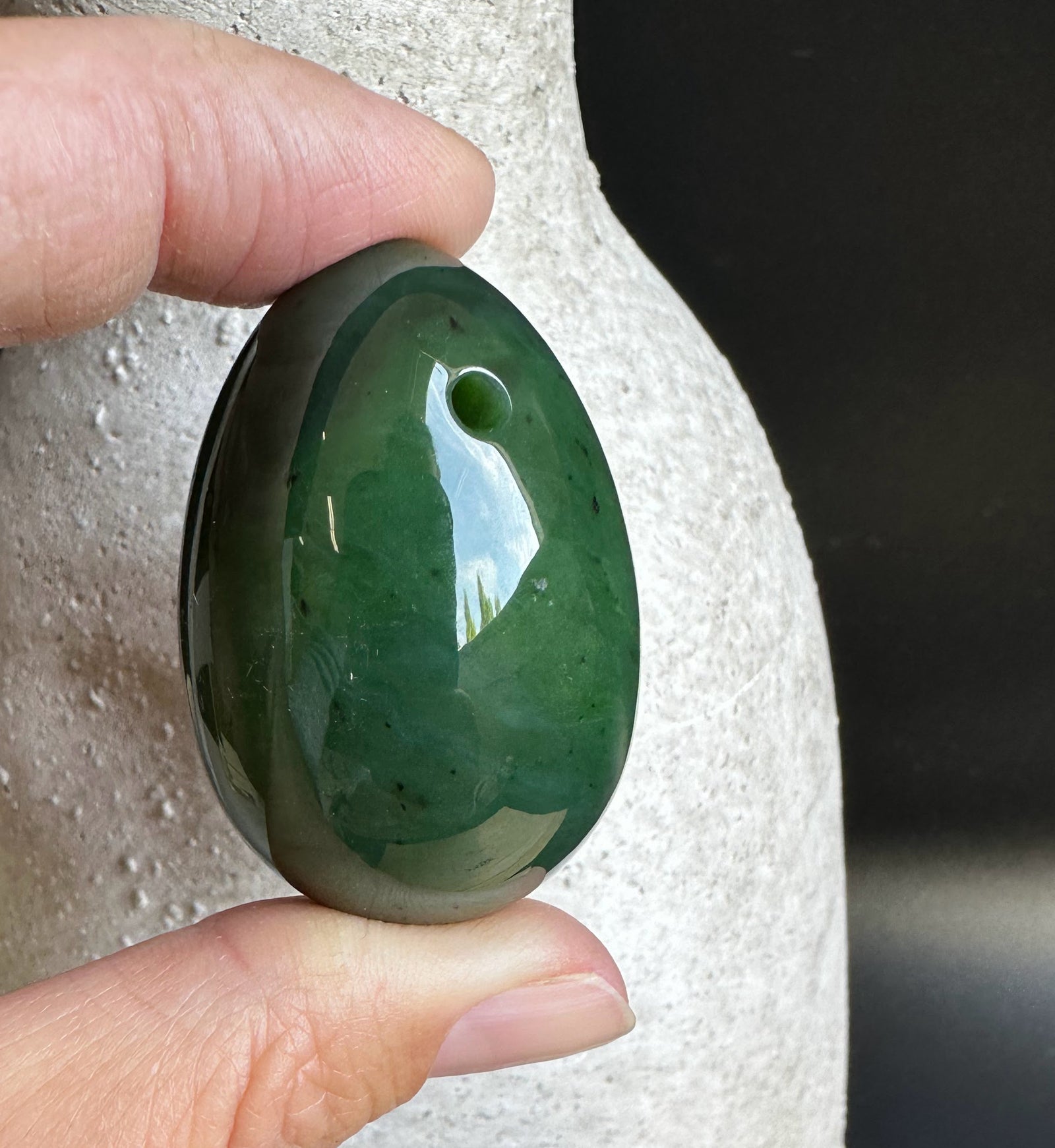 Canadian nephrite yoni egg 1.75 inches held between fingers, glossy dark green stone with a small hole near the rim