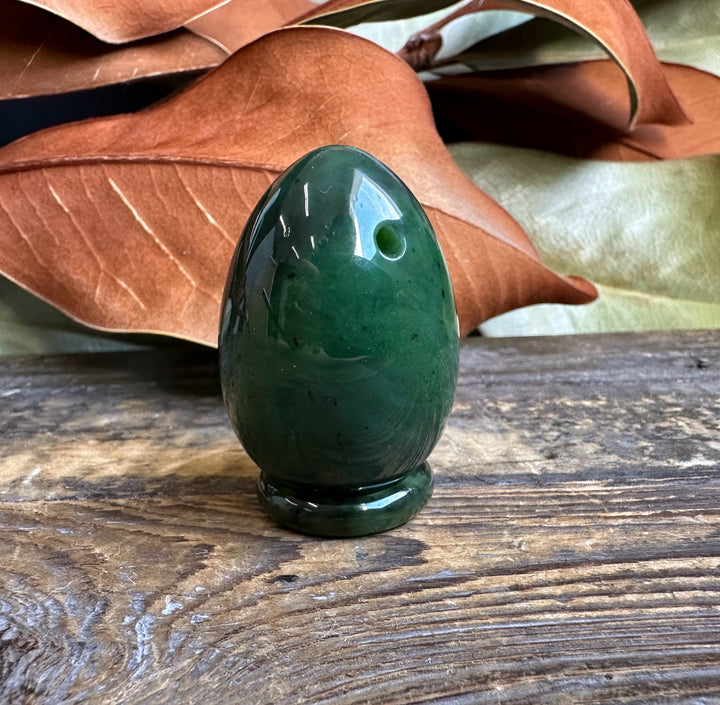 Canadian nephrite yoni egg 1.75 inches, deep green polished stone with a small hole, displayed on a rustic wooden surface.