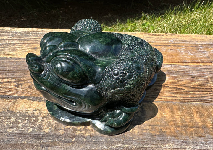 Dark green Ogden Jade Toad sculpture resting on a weathered wooden bench outdoors.