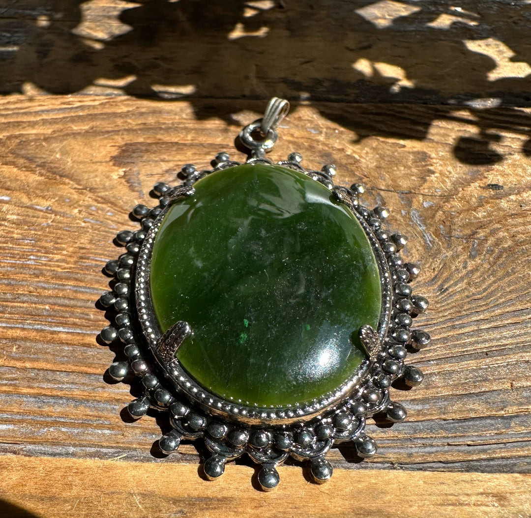 Large vintage jade oval pendant with a silver beaded surround and bail on a wooden surface