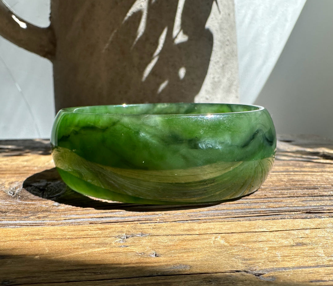 A Grade Ogden jade bangle in marbled green hues rests on a wooden table in sunlight